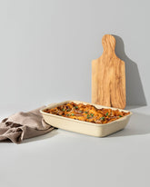 Rectangular beige stoneware baking dish filled with baked pasta, shown on a white surface with a Natural Olive Wood cheese board and a folded fouta towel placed beside it.